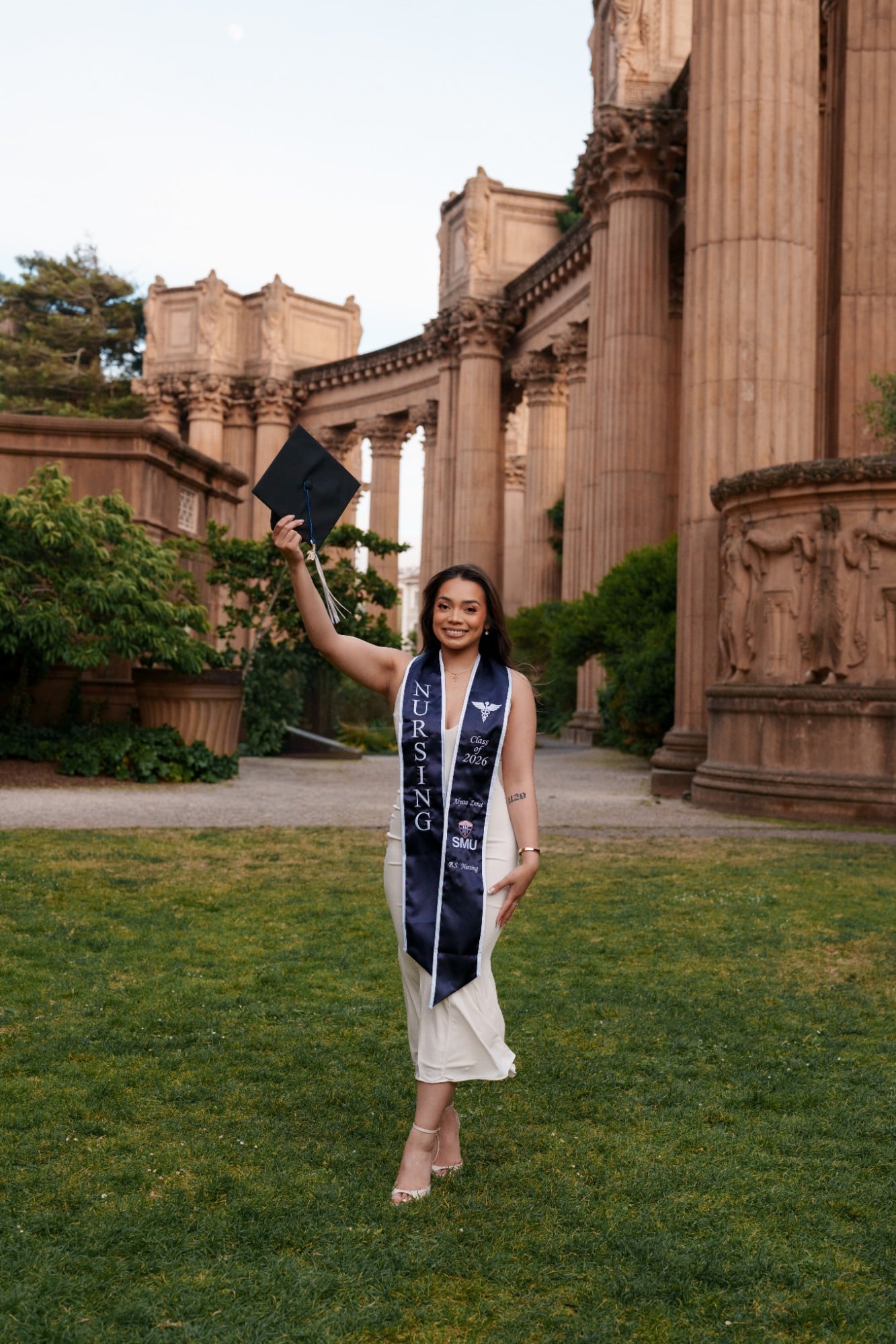 SMU nursing grad in white dress raising her mortarboard beside the Palace of Fine Arts colonnades
