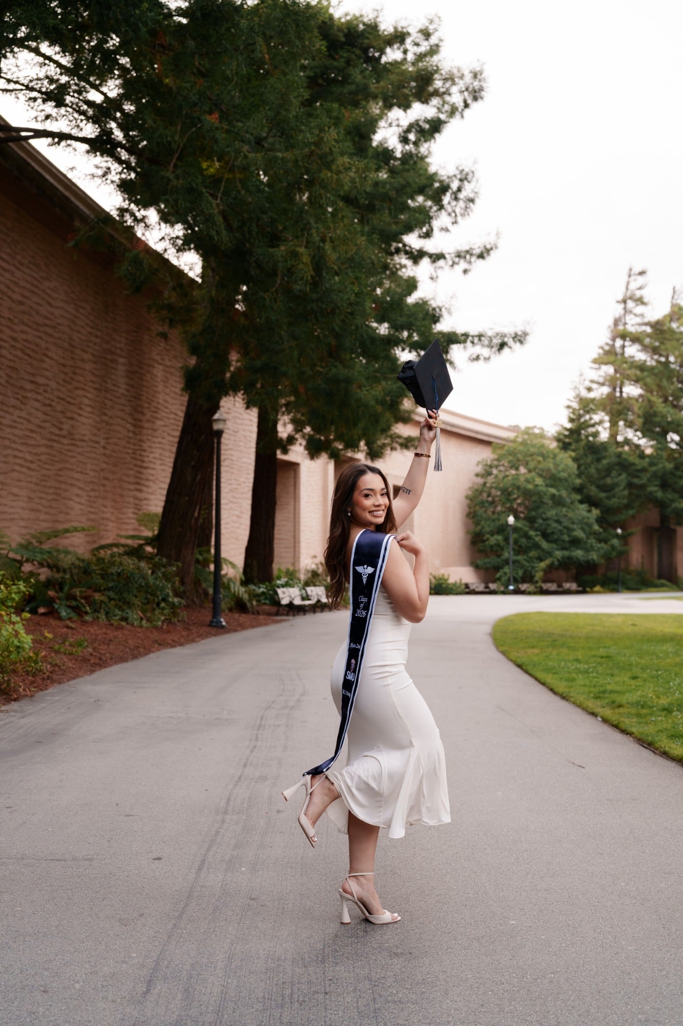 SMU nursing grad in white dress twirling and raising her cap on a tree-lined path near Palace of Fine Arts
