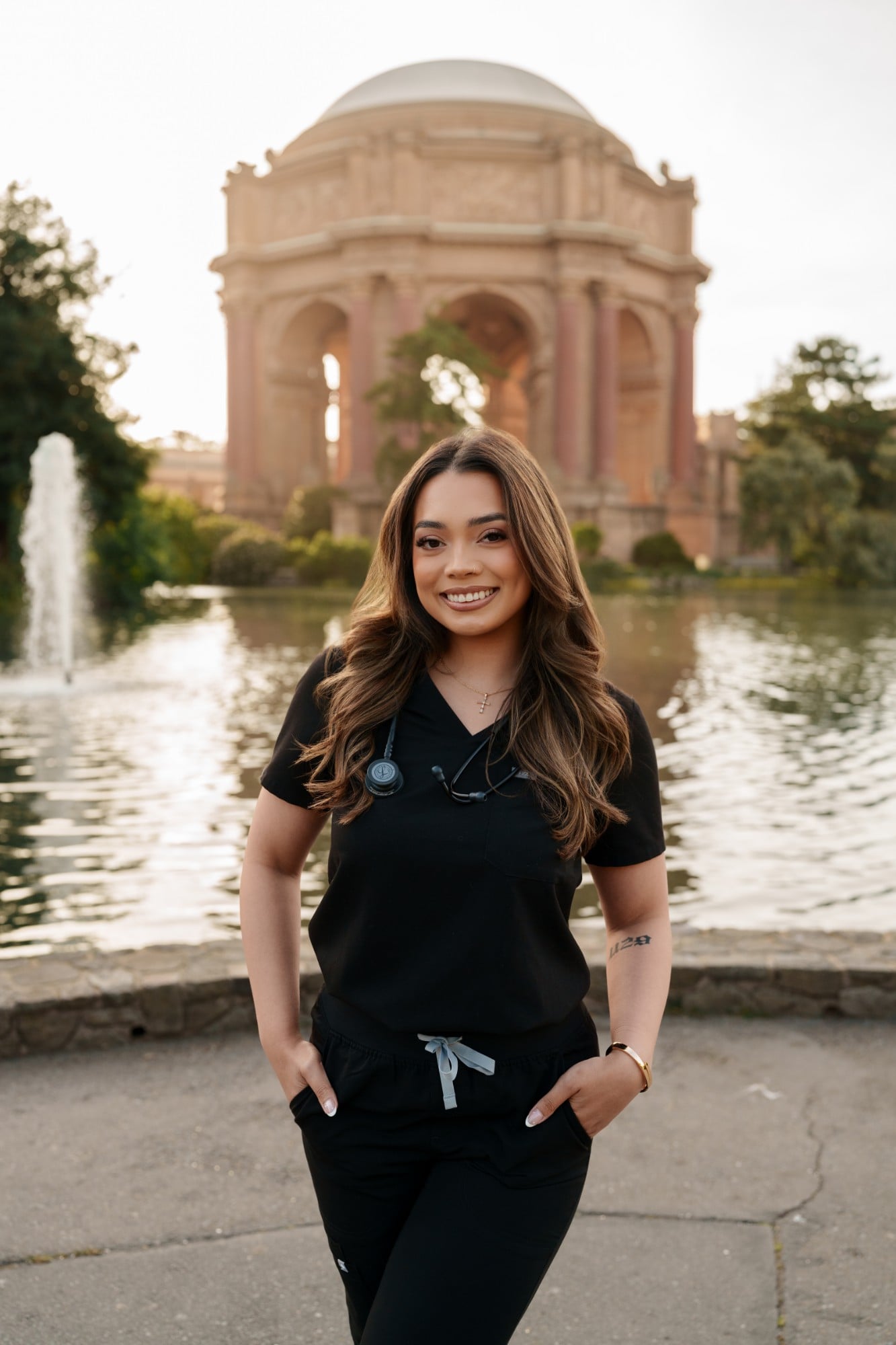 SMU nursing grad in black scrubs with stethoscope hands in pockets smiling at Palace of Fine Arts lagoon