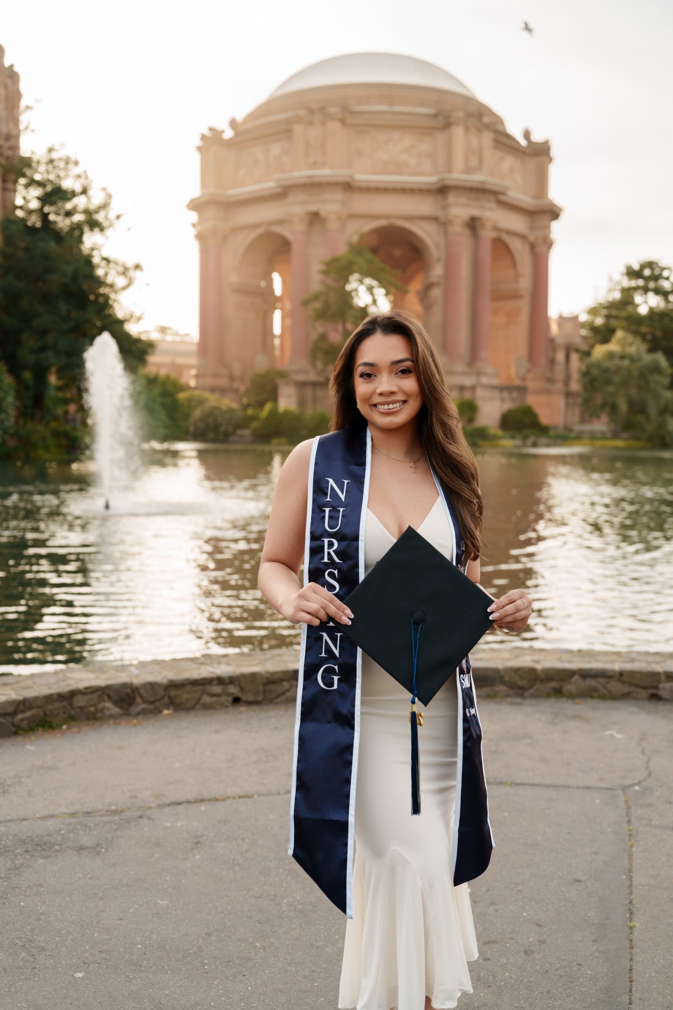 SMU nursing grad in white dress holding cap and navy nursing stole in front of Palace of Fine Arts rotunda and fountain