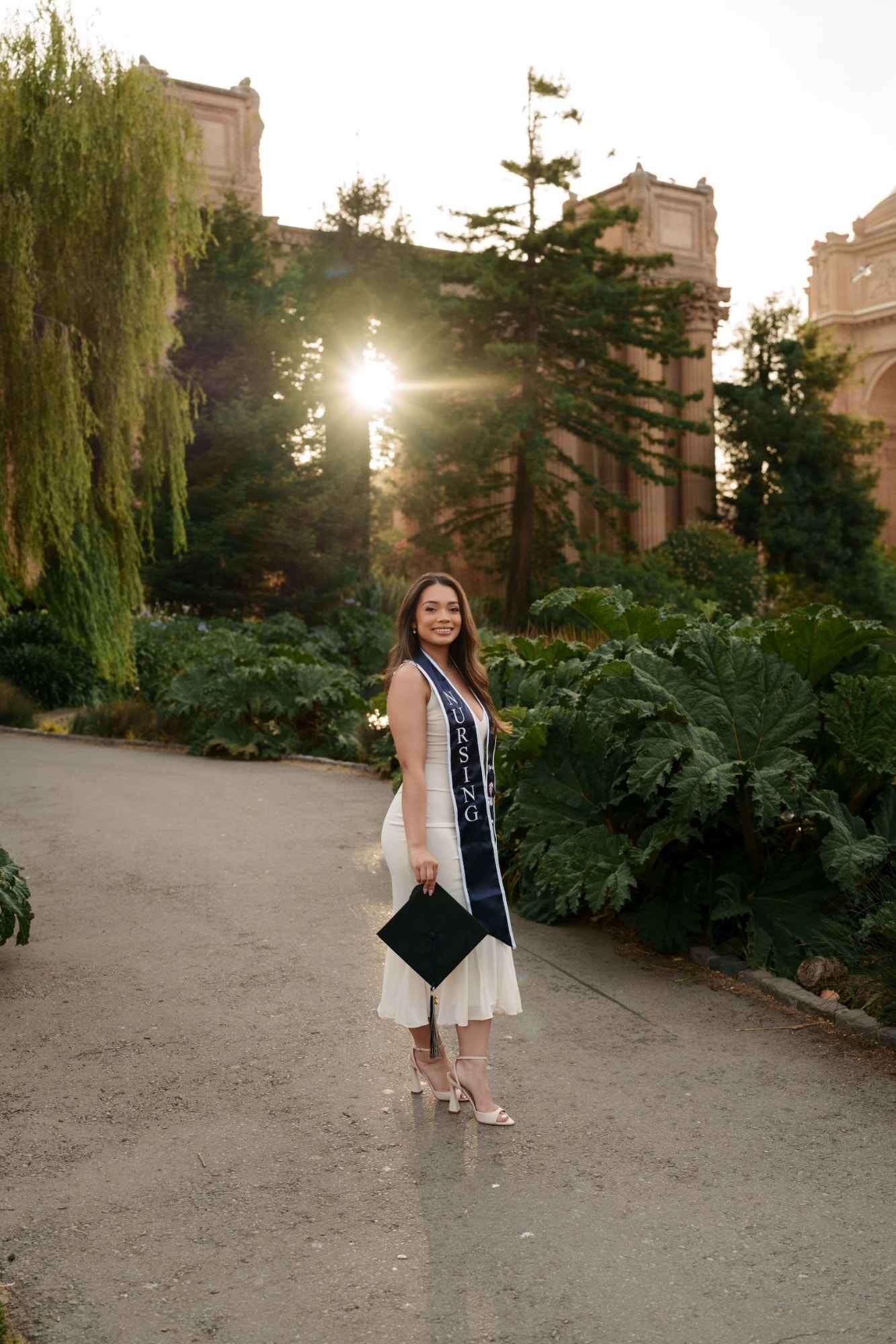 SMU nursing grad in white dress and navy nursing stole holds her cap on a garden path at Palace of Fine Arts at golden hour