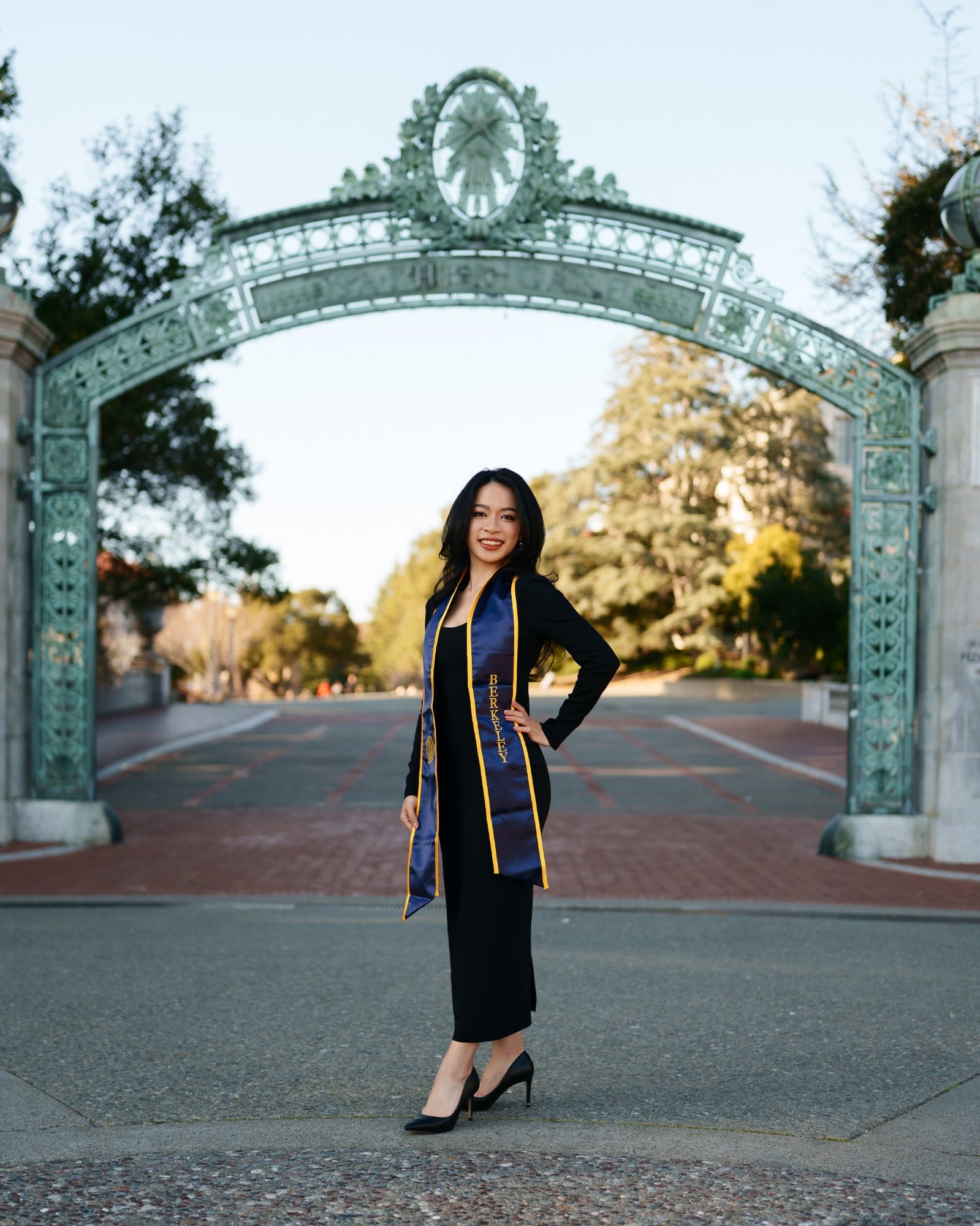 UC Berkeley graduate in black dress and navy-and-gold Berkeley stole standing centered beneath Sather Gate on a clear winter