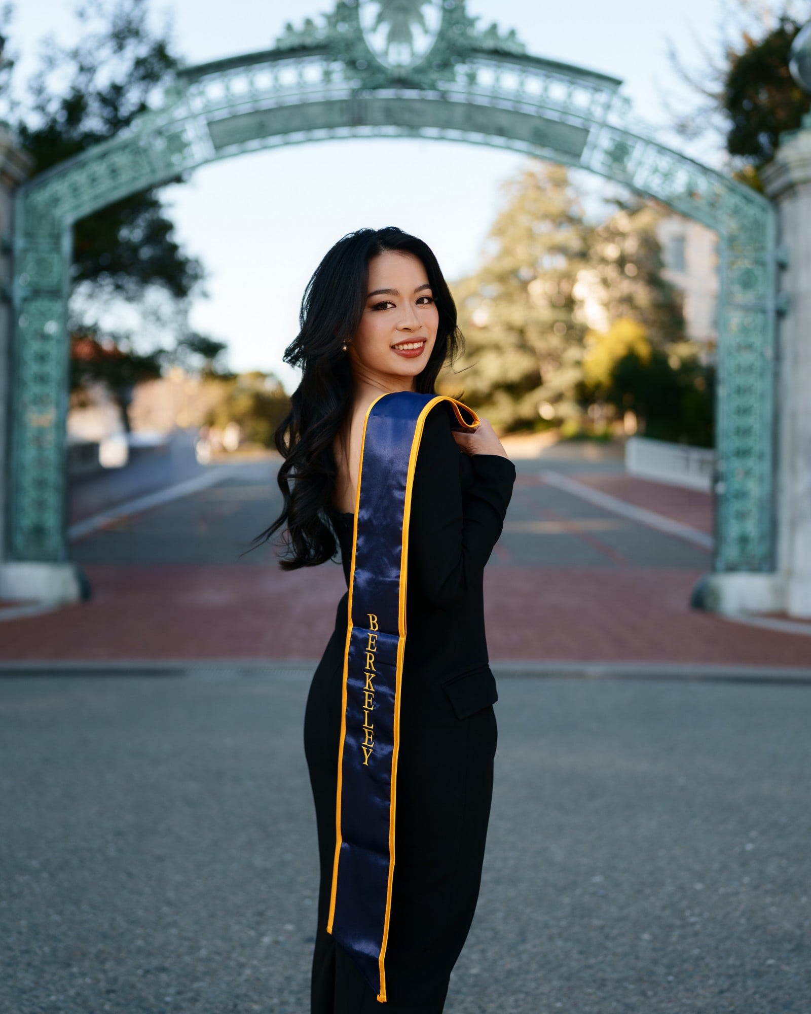 UC Berkeley grad turning over her shoulder at Sather Gate, dark waves catching winter light with navy Berkeley stole draped d