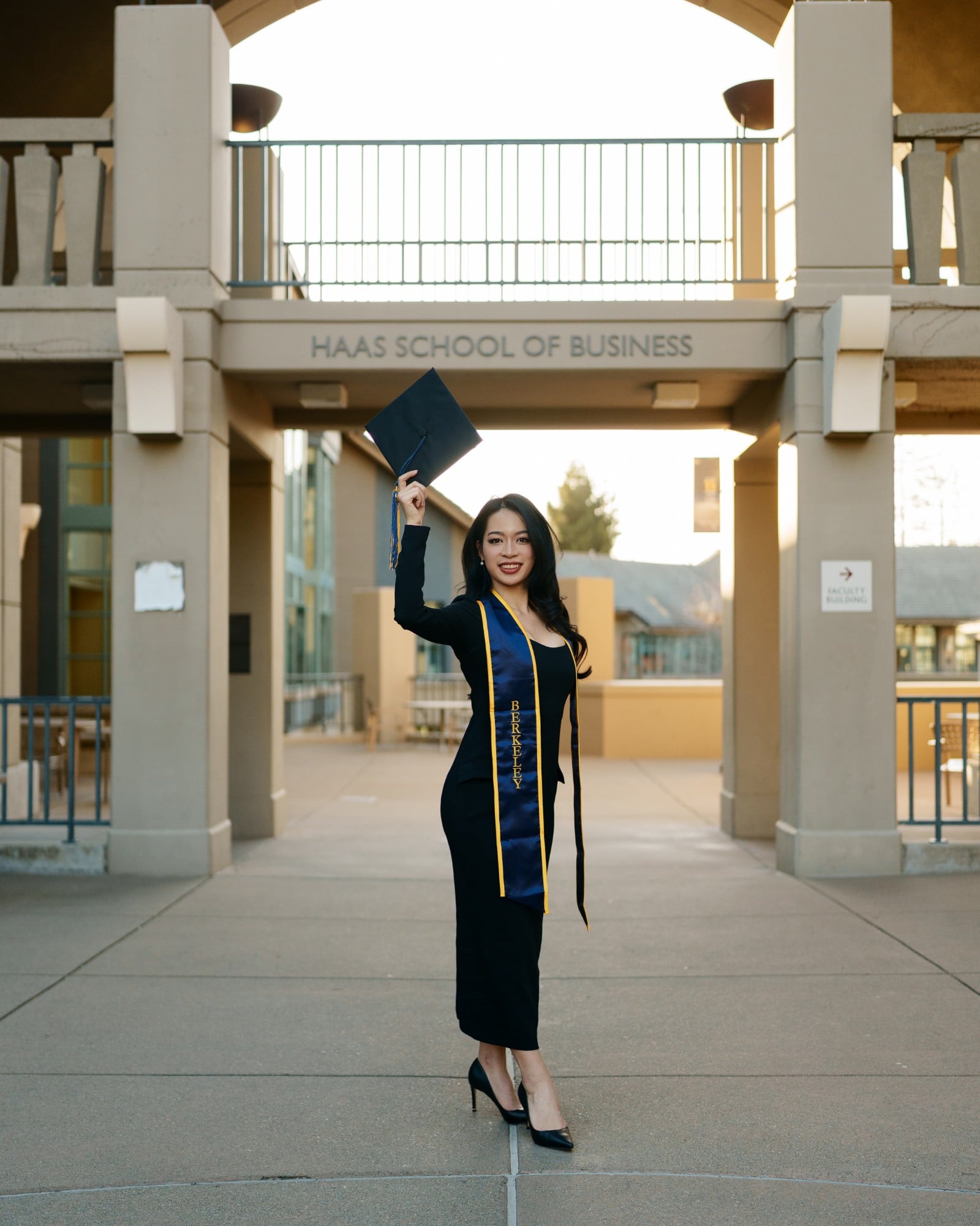 UC Berkeley graduate raising her mortarboard cap and smiling in front of the Haas School of Business entrance sign in warm su