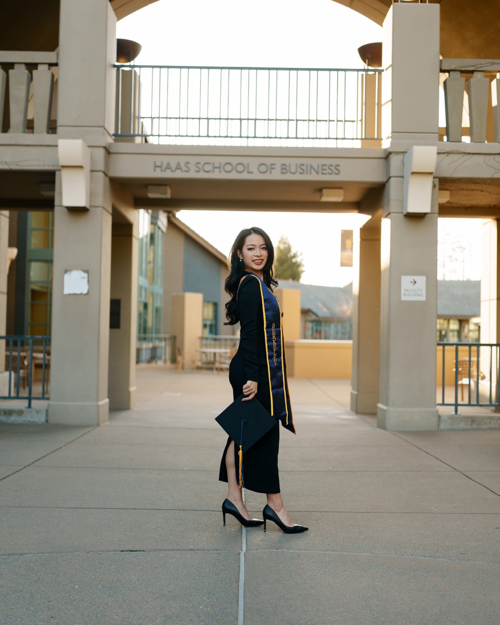 UC Berkeley graduate walking away from camera holding mortarboard at her side at the Haas School of Business courtyard entran