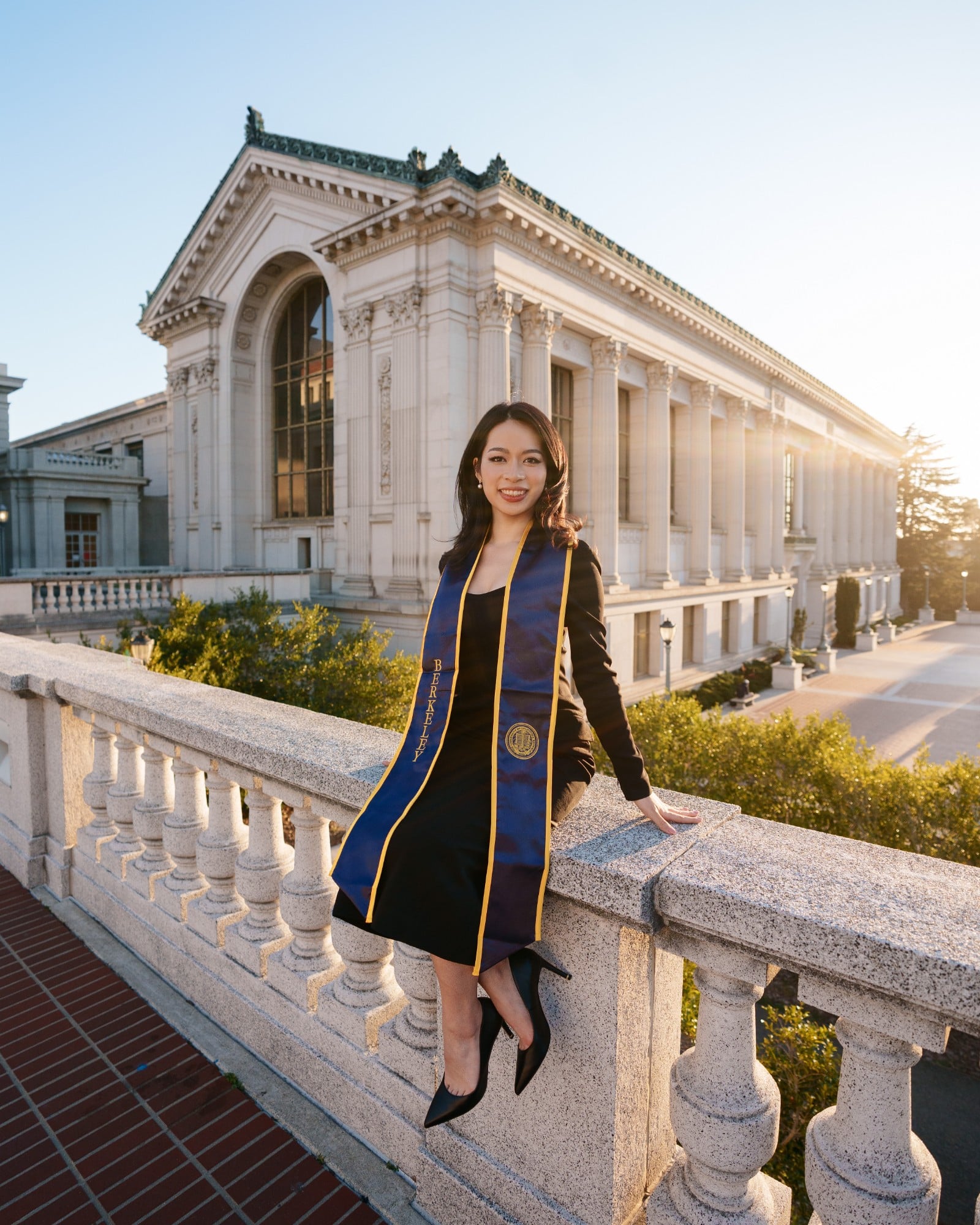 Female UC Berkeley graduate leaning against stone balustrade in front of sunlit Doe Library columns wearing navy-and-gold Ber