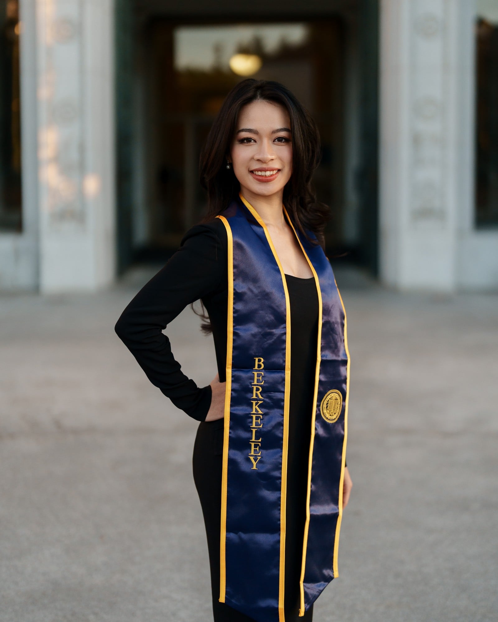Close-up portrait of UC Berkeley graduate smiling with navy-and-gold Berkeley stole and gold seal, ornate Doe Library entranc