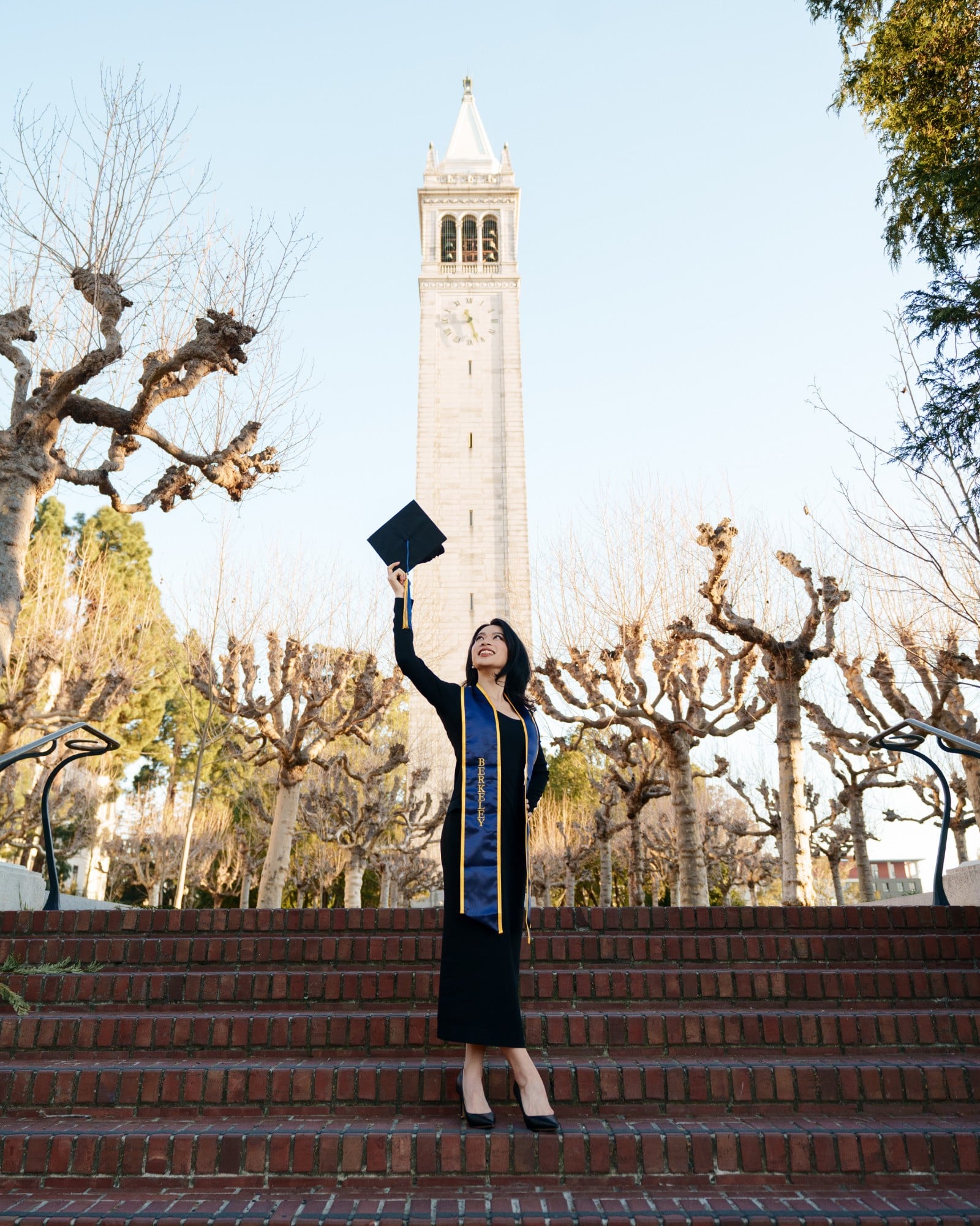 UC Berkeley graduate raising her mortarboard cap on brick steps below the Campanile clock tower, bare winter trees framing th