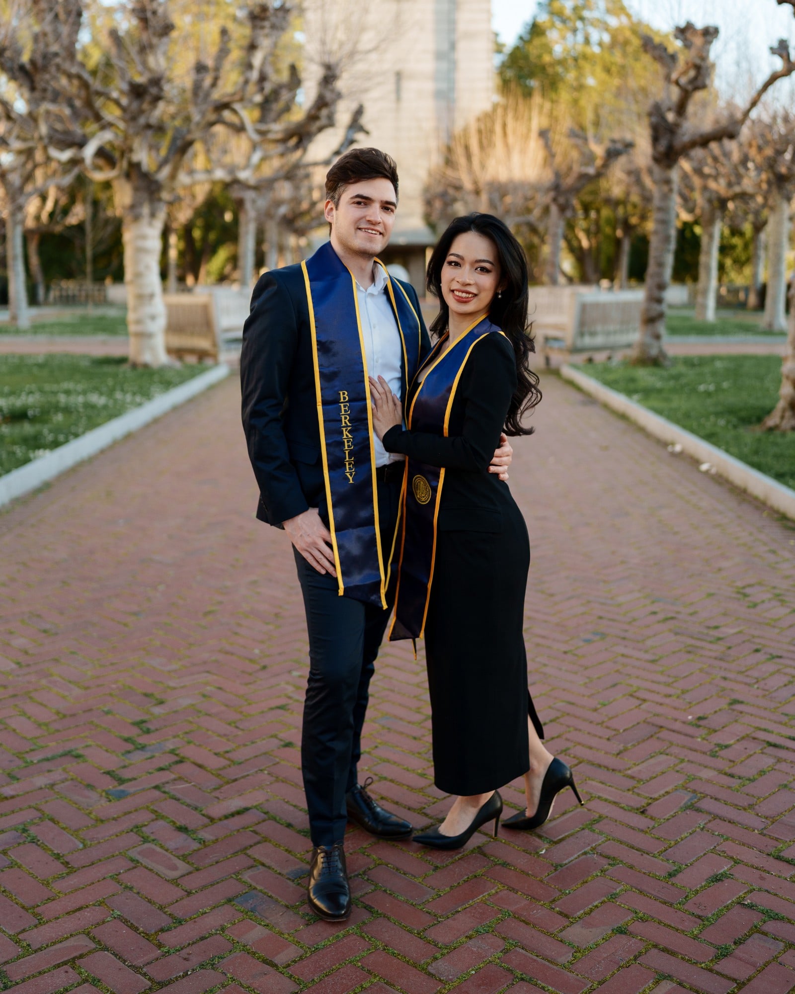 Two UC Berkeley graduates in matching navy-and-gold Berkeley stoles embracing on a brick walkway lined with pruned sycamore t