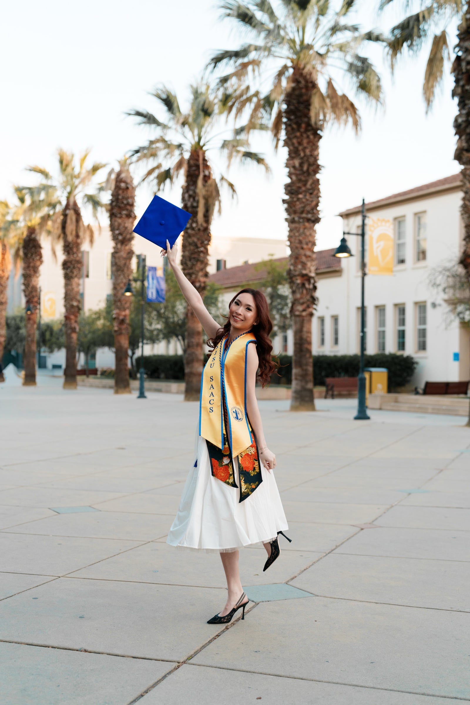 SJSU graduate tossing her blue cap among palm trees on San Jose State's main walkway wearing a white midi dress and gold SAAC